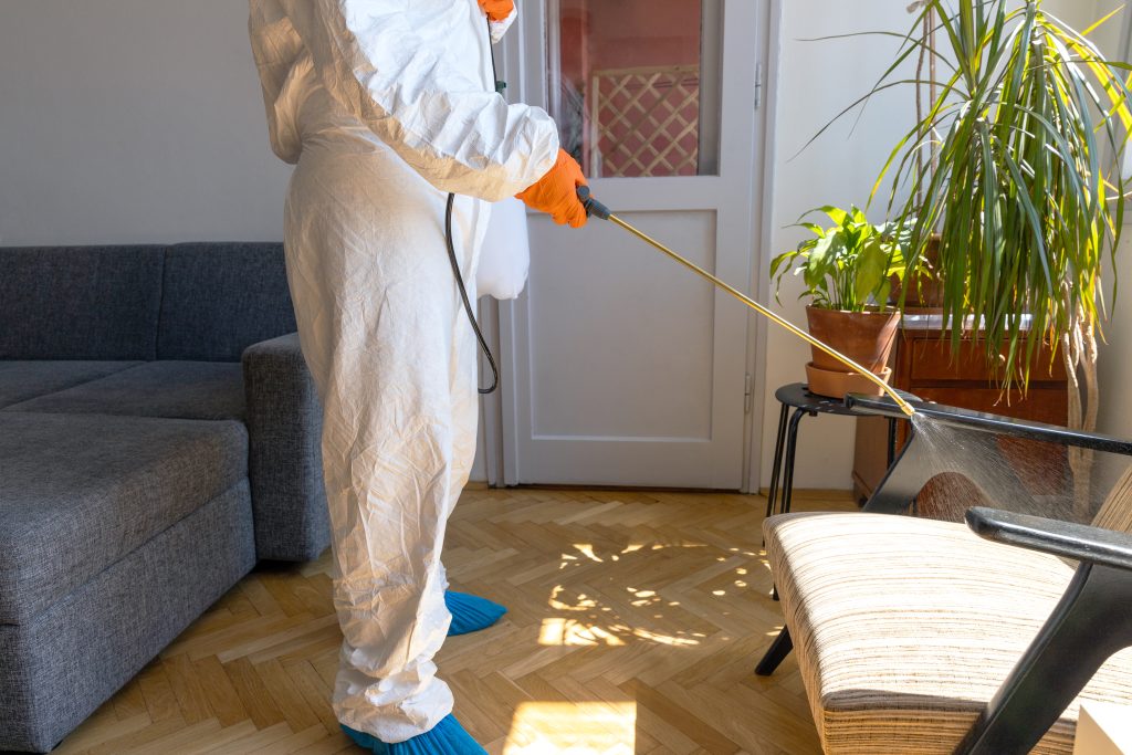 Worker in protective suit with decontamination sprayer bottle disinfecting household and furniture. Pest control concept.