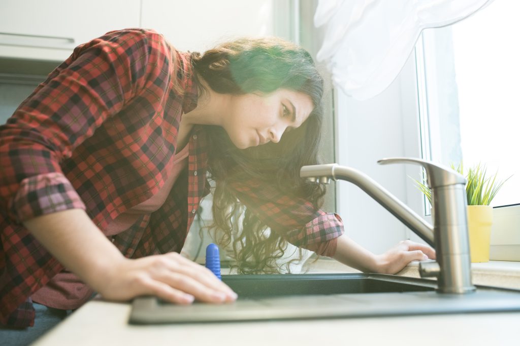 Serious concentrated young woman in checkered shirt checking faucet while having problem with dropping faucet in kitchen