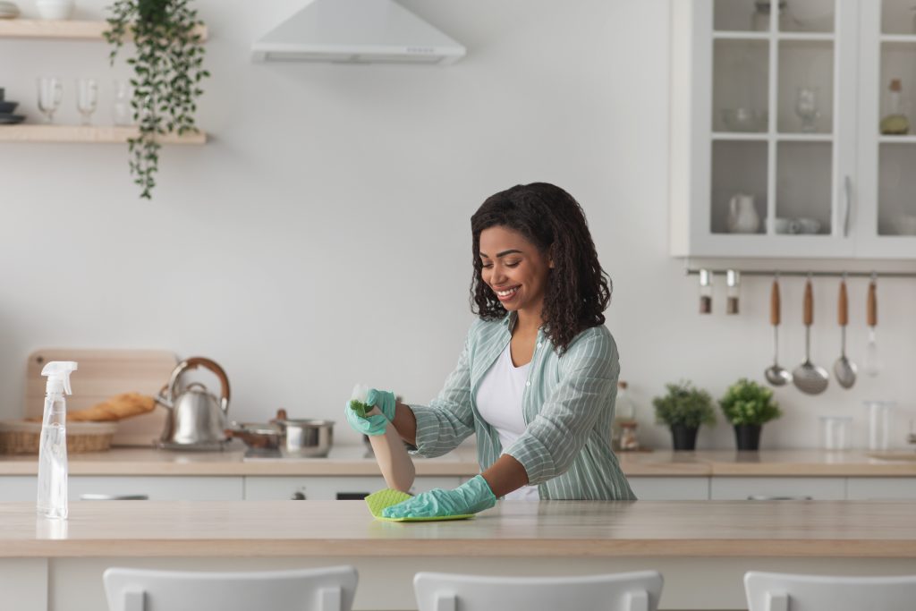 Housewife, daily home cleaning, routine use napkin wipe dust and mud from table or desk, housekeeping. Smiling young african american woman in protective gloves with sponge and spray in white kitchen