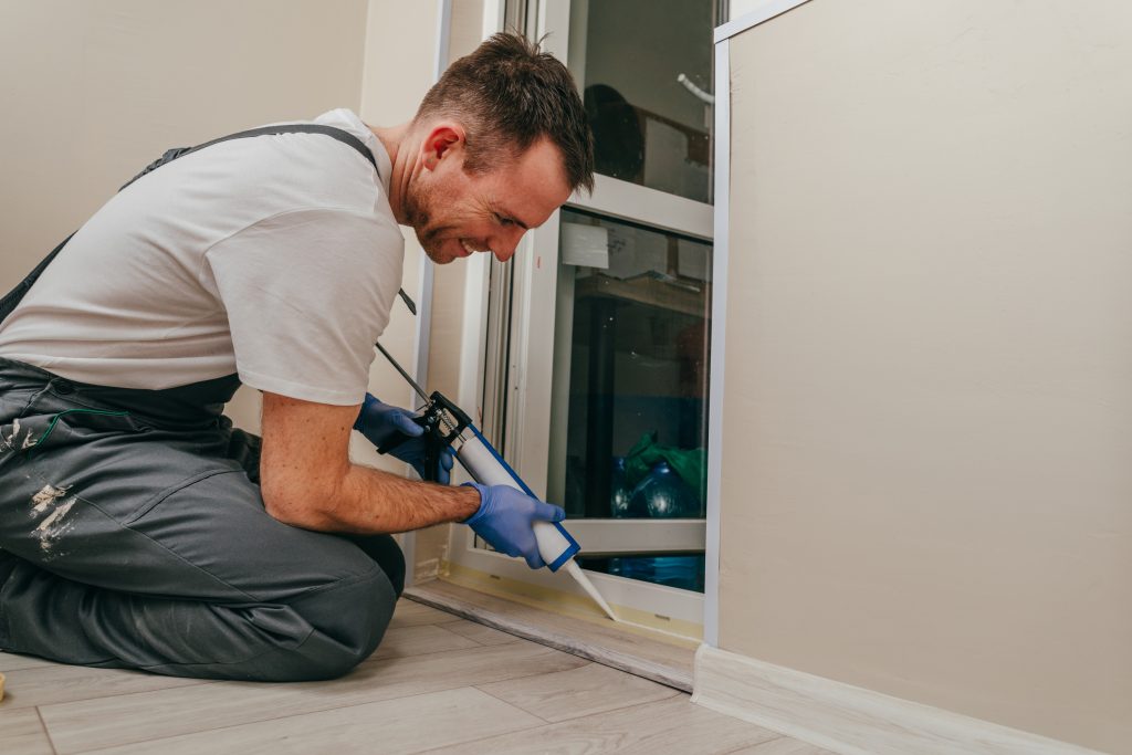 Young man wearing overalls sealing cracks between door and trim using waterproof silicone caulk on the balcony.