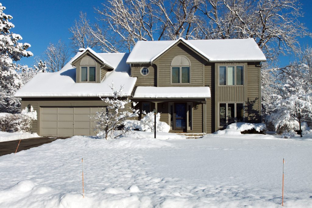 Color DSLR stock picture of green and grey suburban colonial style house covered in bright, white winter snow. Clear, cold background for home is bright blue sky. Black top driveway is clear, but lawn is blanketed. No people in shot but ample copy space for text